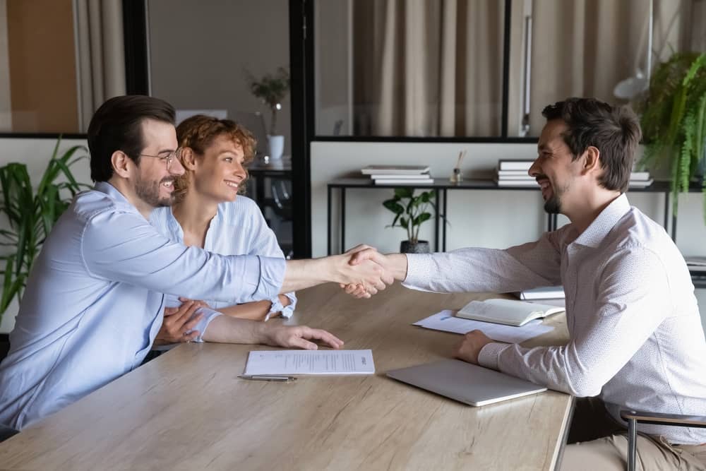 Confident lawyer meeting and shaking hands with happy couple of clients. Husband and wife visiting office of mortgage broker, real estate agent, house seller shaking hands over signed contract