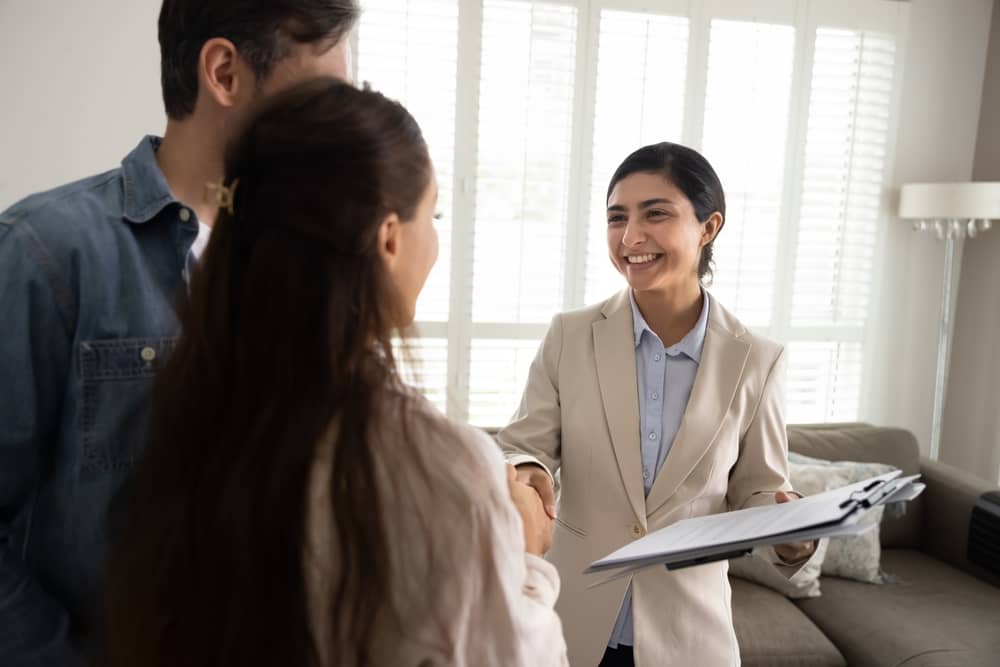 Happy young Indian real estate agent meeting with couple of house buyers indoors, shaking hands after signing agreement, holding legal paper documents, smiling, congratulating on purchase
