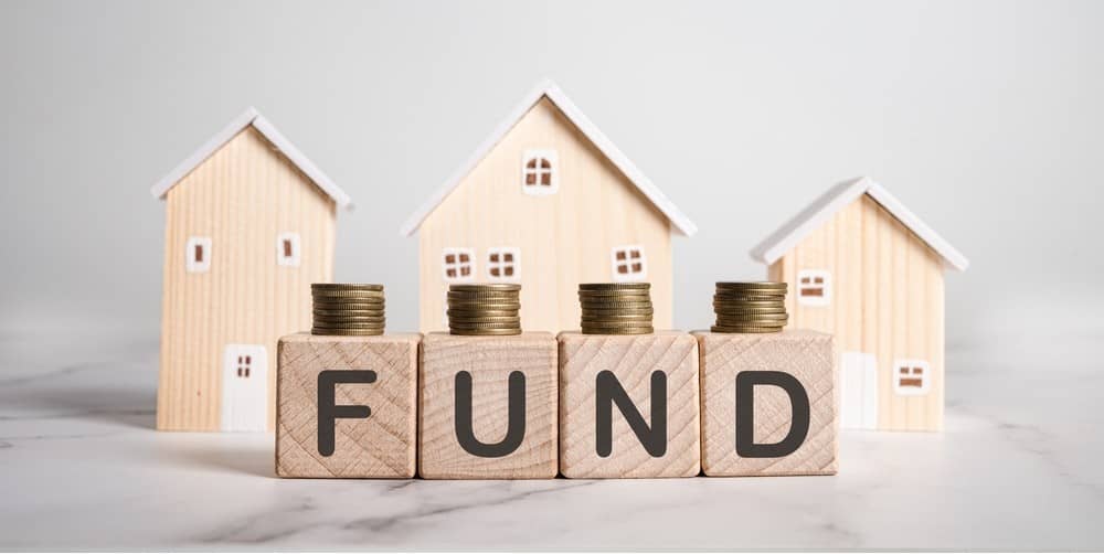Wooden blocks spelling FUND topped with coin stacks in front of miniature houses. Property investment funds, home savings, mortgage planning, and real estate asset management.