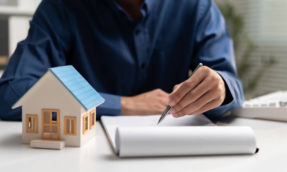 Real estate businessman reviews property documents beside a house model, representing home purchase, mortgage, contract, property investment, financing, ownership, and housing agreement.